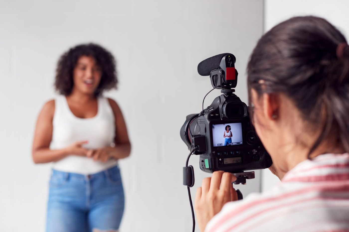 Woman with camera recording female vlogger in white top and blue jeans.