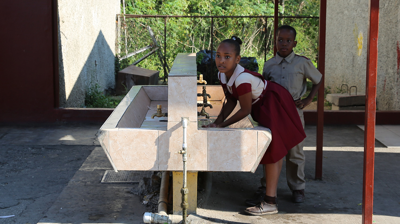 A schoolgirl in red and white school uniform is washing hands at an outside lavatory while a schoolboy in sand uniform is waiting behind her