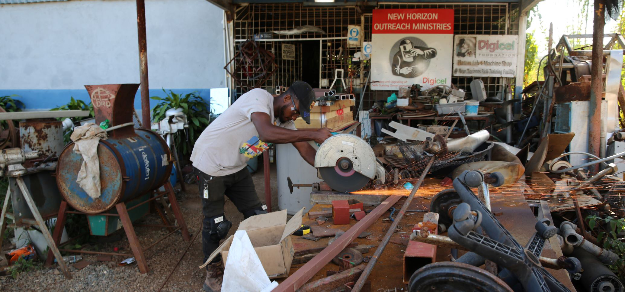 A trainee wearing a black baseball cap and trousers, a sand T-shirt, and protective goggles is cutting a piece of metal with a disc saw