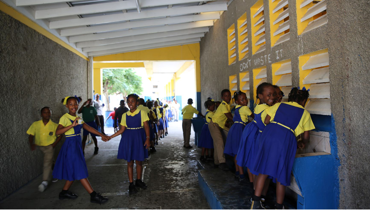 Female students, wearing yellow shirts and blue skirts, and male students, wearing yellow shirts and kaki trousers, are walking through a covered passageway with lavatories