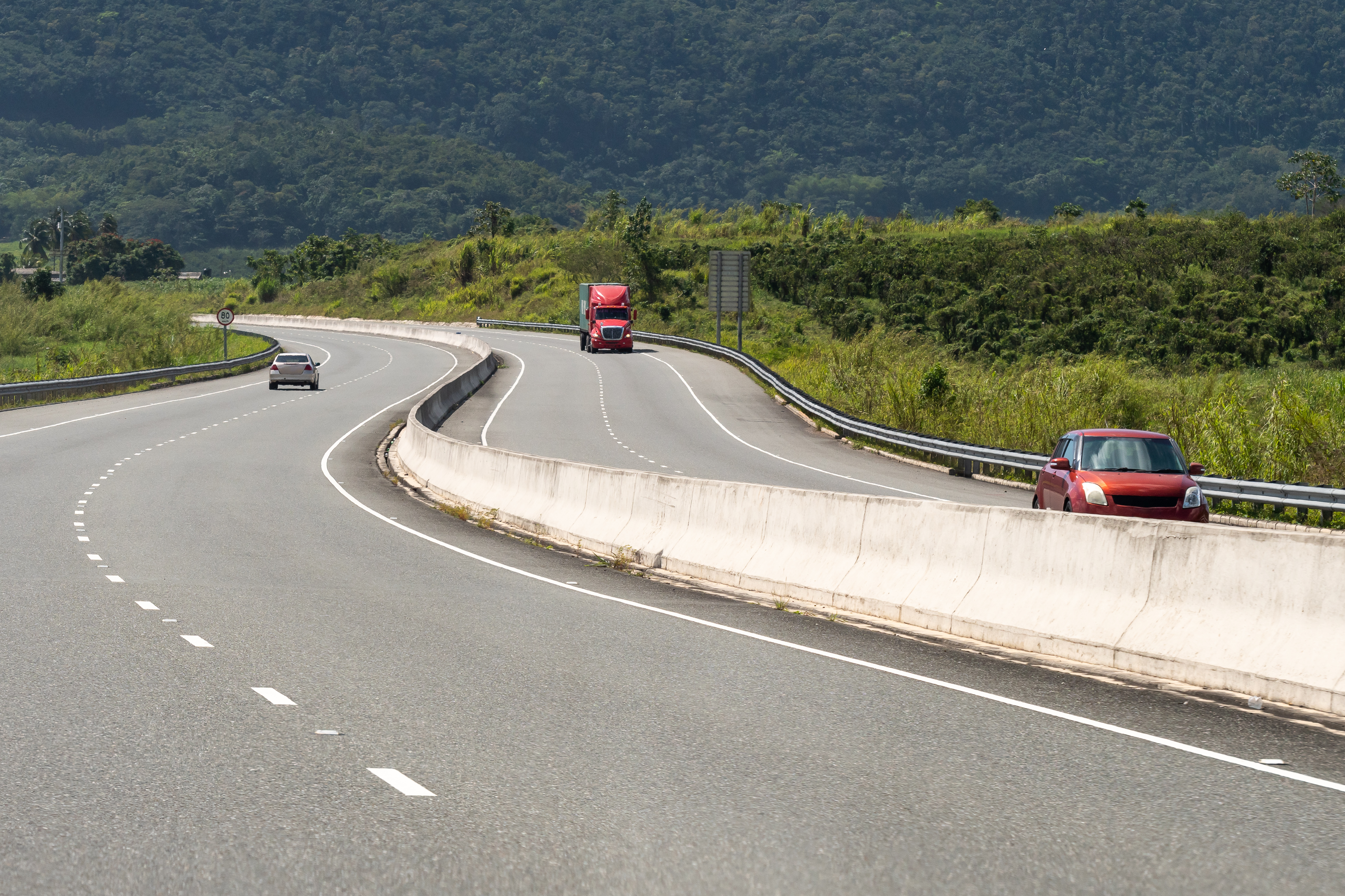 Dual/ double/ two lane carriageway highway through scenic countryside mountains. Cars, trucks, vehicles drive on left hand side of the street. Winding road in Saint Ann/St Ann, Jamaica.