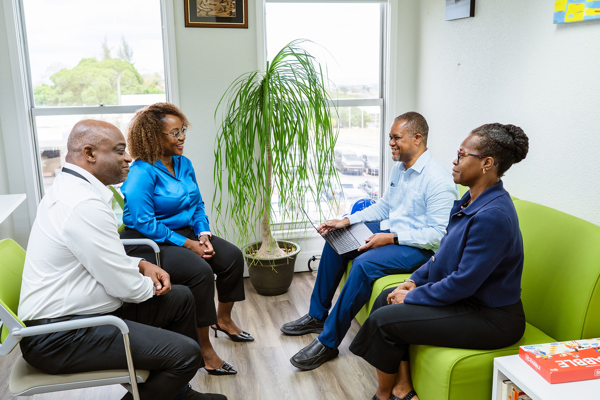 Four professionals seated in a small office meeting, with a laptop and a potted plant by a window.