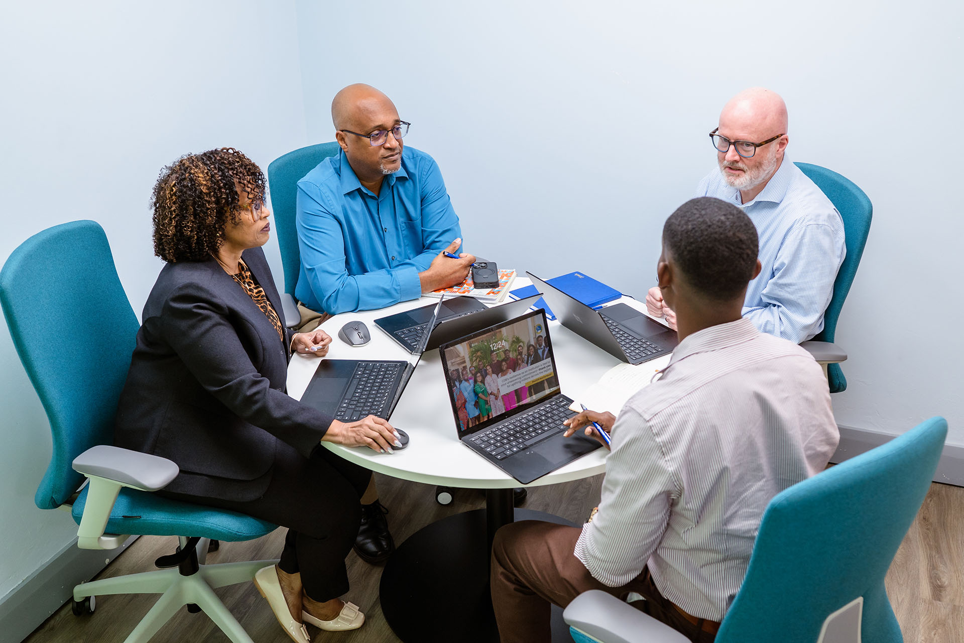 Four professionals seated around a table in a meeting, using laptops and discussing work.