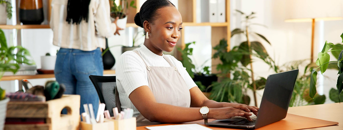 Person working on a laptop at a desk in a bright, plant-filled workspace with shelves and stationery in the background.