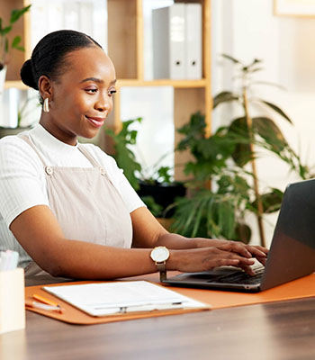 Person working on a laptop at a desk in a bright, plant-filled workspace with shelves and stationery in the background.