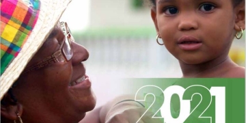 afro Caribbean grandmother in a read shirt and colourful hat holds a female toddler in her arms as toddler looks into the camera