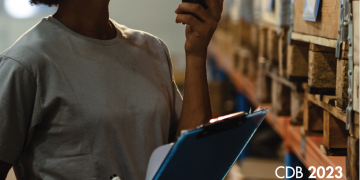 Black woman in grey shirt and blue pants with walkie talkie and clipboard looking up at shelves 