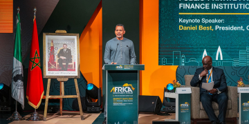 President Best standing at a lectern delivering remarks at the Africa Investment Forum 2025. A framed photograph of a Moroccan dignitary is displayed on an easel to the left, and event branding is visible on the stage backdrop.
