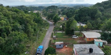 Aerial view of a rural road winding through a green, hilly landscape with scattered houses and vehicles, showing community infrastructure in a tropical area