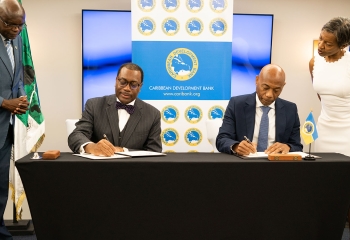 two seated gentlemen signing document as others look on