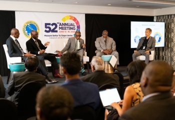 Five panellists seated on dark riser with multicoloured logo on screen behind them.