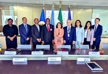 group photo with males and females standing behind a large table and flags in the background