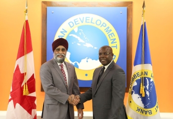 Minister Harjit Sajjan shaking hands with CDB Vice President Isaac Solomon in front of flags of Canada and CDB