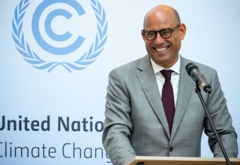 a smiling simon stiell wearing grey suit standing at lectern