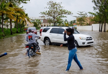 Woman walks across flooded street covering her head. A man is riding a bike, and a car is driving through the flooded streets. ed streets.