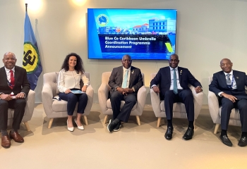 Five individuals are seated in a room below a television screen with a CARICOM flag to the right 