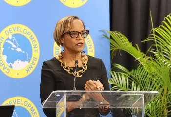 black woman waring glasses and a black dress standing at acrylic lectern with step and repeat backdrop behind her