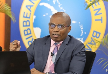 black man wearing a grey suit with pink shirt and tie seated at a desk with an open laptop