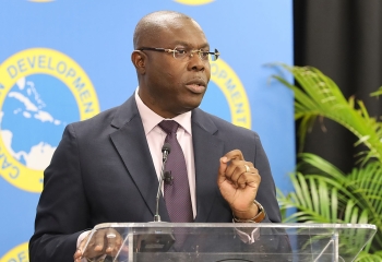 black man wearing glasses in dark suit with pink shirt and mauve tie standards at acrylic lectern delivering remarks