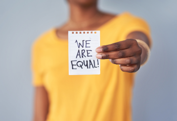 woman in yellow shirt holding hand outstretched with paper with the words 'We Are Equal'