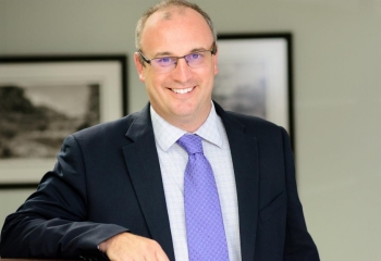Caucasian male wearing glasses dressed in black suit with lilac tie looks into camera while smiling