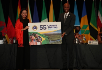 A woman and man stand on a platform in front of several flags of CDB member countries. The man is handing over a card to the woman with the words 55th Annual Meeting 