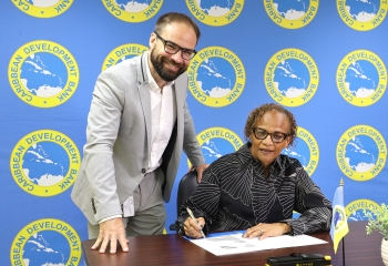 Lady seated in black and white patterned with pen signing and man in tan suit leaning over table and looking forward
