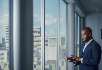 Thoughtful Black Businessman in a Tailored Suit Using Laptop while Standing in Office Near Window on Big City.