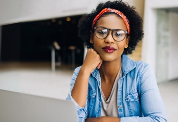 A confident woman with natural curly hair wearing glasses and a red patterned headband sits at a desk. She rests her chin on her hand while looking directly at the camera. She wears a light denim jacket over a beige top. The background features a softly lit modern interior with blurred details.