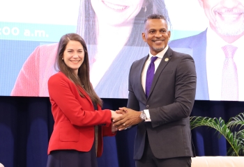 A woman and a man in formal attire are shaking hands and smiling for the camera against a stage backdrop, with their images displayed on a screen behind them