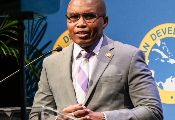 CDB Director of Economics, Mr. Ian Durant dressed in a grey suit with a pink and grey tie speaks at a podium in front of a backdrop displaying the logo of the Caribbean Development Bank.