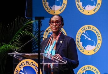 CDB Division Chief, Environmental Sustainability,, Ms. Valerie Isaac dressed in adark suit with colourful inside shirt speaks at a podium in front of a backdrop displaying the logo of the Caribbean Development Bank.