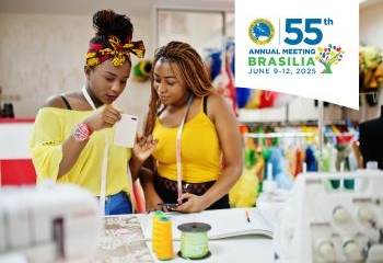 The image shows two women in a brightly lit fashion workshop. They are focused on a smartphone, seemingly discussing something related to their work. Both are wearing vibrant yellow outfits, with measuring tapes around their necks, and are surrounded by colorful fabric designs and sewing equipment in the background.