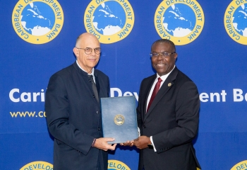 Two men in formal attire standing front of a blue backprop with the Caribbean Development Bank logo. They are smiling and holding a blue folder embossed with the Bank’s seal, during the signing ceremony.