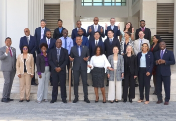 A diverse group of professionals stands together on the steps outside a modern building for a group photo. The individuals are smiling, gesturing positively, and dressed in formal business attire, indicating a celebratory or official event. The setting suggests a conference or institutional gathering, with architectural columns and large windows in the background.