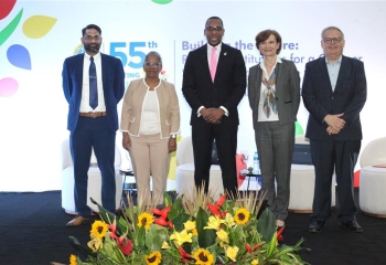 **Description for Accessibility:** A group of five individuals stands on a stage at the 55th Annual Meeting of the Caribbean Development Bank, with the event's theme "Building the Future: Resilient Institutions for a Greener, Stronger, and Inclusive Caribbean" displayed on the backdrop. The individuals, representing a mix of formal attire, are positioned in front of a vibrant floral arrangement featuring sunflowers and tropical plants. The event logo is prominently visible in the background.