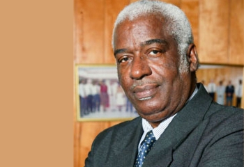 Corporate portrait of a grey haired elderly black man in a black business suit against a brown background