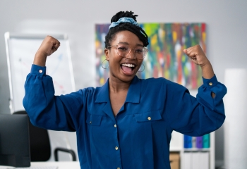 Dark-skinned woman wearing blue clothes glasses stands against office backdrop smiles happy, victory, winning, raises hands up tenses hand muscles gestures strength power successful girl businesswoman