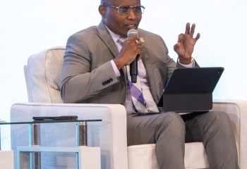 Bespectacled black man seated in a grey suit on a white chair on stage with a mic in hand gesticulating and looking ahead 
