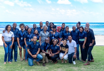 A large group of black entrepreneurs standing and stooping in navy blue tshirts on grass under a clear blue sky and ocean