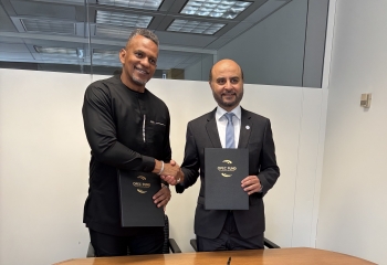 two men in business attire standing in a room behind a table holding up branded folders and shaking hands 
