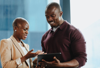 Black man and woman dressed professionally looking down at a tablet and smiling 