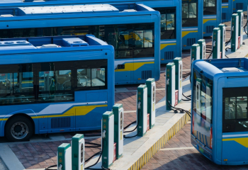 Row of blue electric buses parked at a charging station with multiple charging units on a paved lot.