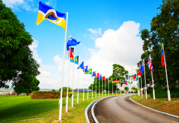 Row of Caribbean Development Bank member country flags lining a curved driveway outside the CDB headquarters, waving against a bright blue sky with scattered clouds.
