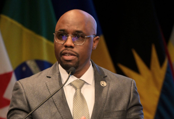 Male speaker at podium addressing audience, with Caribbean flags in background.