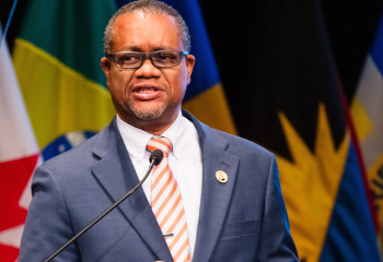 Male speaker in dark blue suit and orange and grey tie at a podium addressing an audience, with multiple national flags visible in the background.