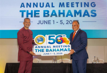 Chairman Halkitis and President Best holding a sign for the Caribbean Development Bank’s 56th Annual Meeting in The Bahamas, with event dates displayed on a stage.