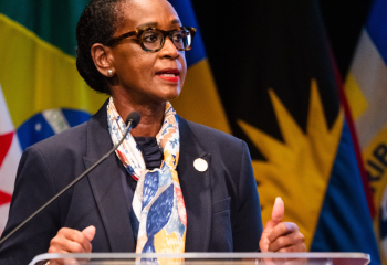 Speaker at a podium addressing an audience, with multiple national flags visible in the background.