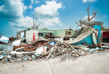Severely damaged buildings and debris scattered across a coastal area after a storm, with collapsed roofs and broken structures under a partly cloudy sky.
