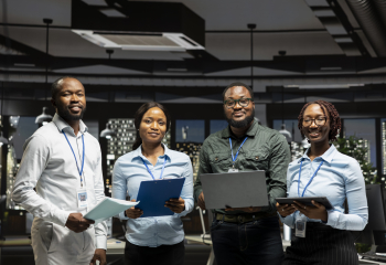 Four professionals standing in a modern office, holding documents, a laptop, and a tablet